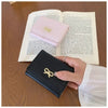 Cartera de cuero PU negra plegable con hebilla decorativa dorada sobre una mesa de madera junto a un libro abierto.