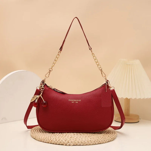 A maroon leather shoulder bag with a gold chain and cherry pendant resting on a woven mat.