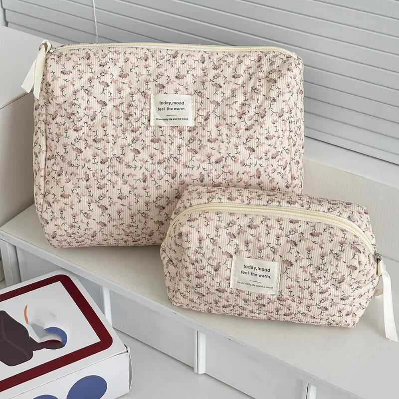 A set of two floral print quilted cosmetic bags with light beige fabric and white trim resting on a white shelf.