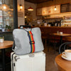 A grey fabric luggage strap with colorful stripes rests on top of a white suitcase in a cafe setting.