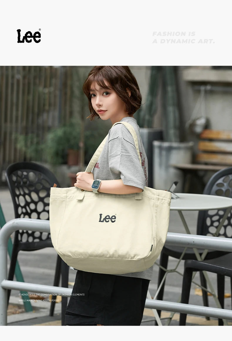 A woman models a beige Lee oxford cloth tote bag in an outdoor setting.