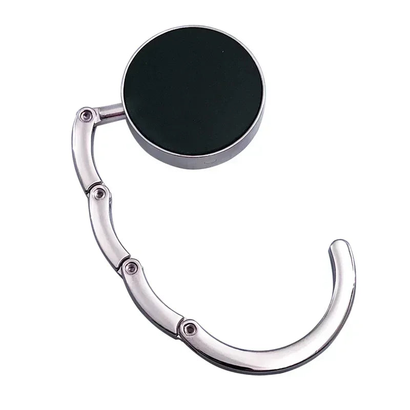 Gancho de bolsa plegable plateada con una placa negra redonda para organizar mesas y bolsos