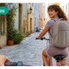A woman wearing a large capacity laptop backpack is walking down a cobblestone street surrounded by greenery.