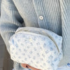 A white makeup bag with light blue stripes and a flip-top mirror is held by a person wearing a light gray knitted sweater.