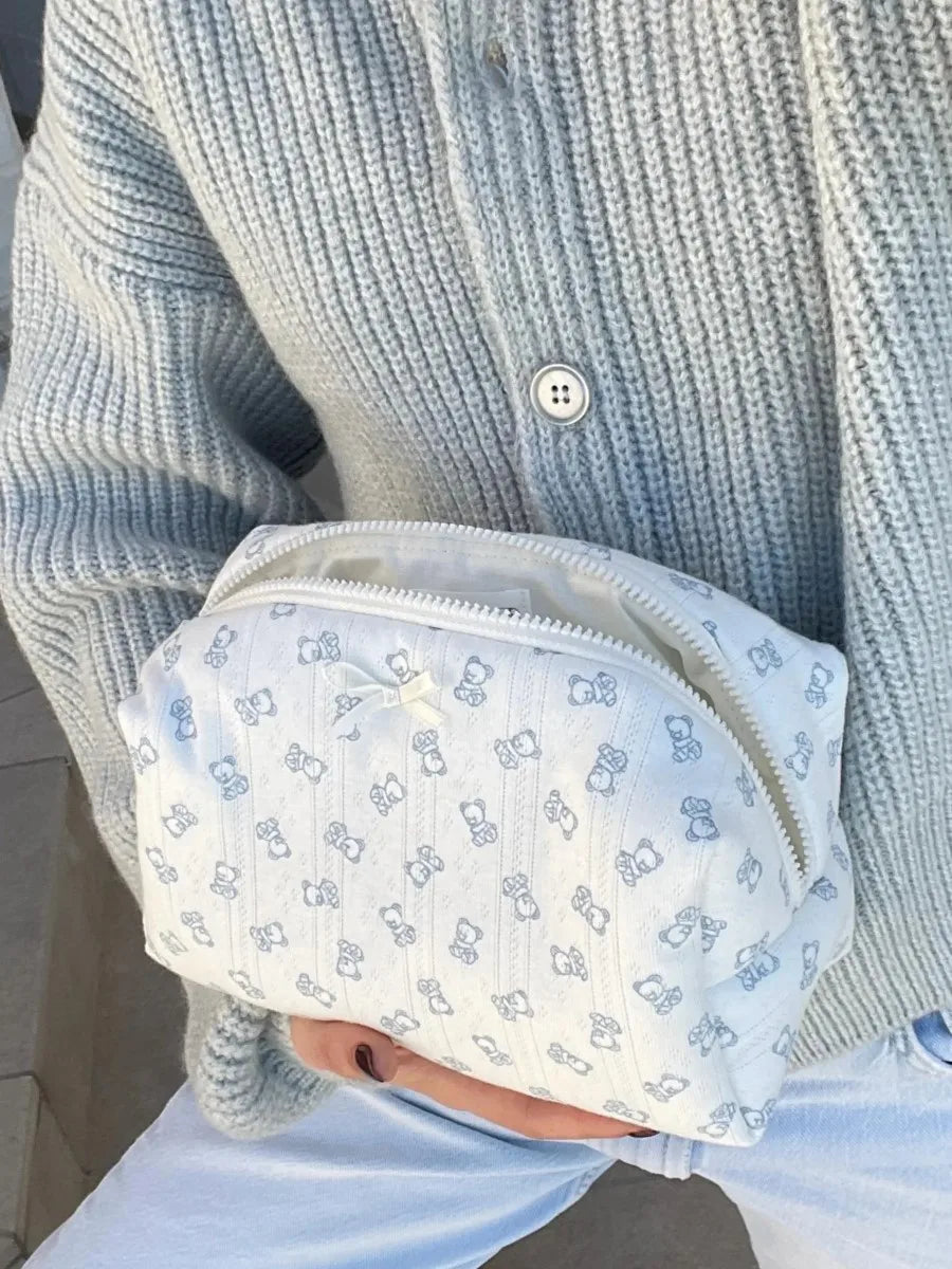 A white makeup bag with light blue stripes and a flip-top mirror is held by a person wearing a light gray knitted sweater.