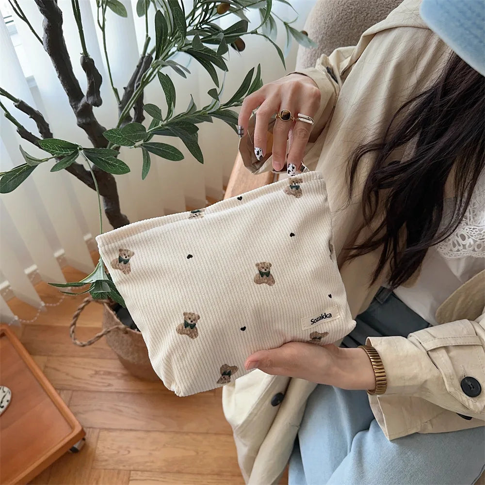 A beige corduroy clutch bag is held by a person, set against a bright indoor background.
