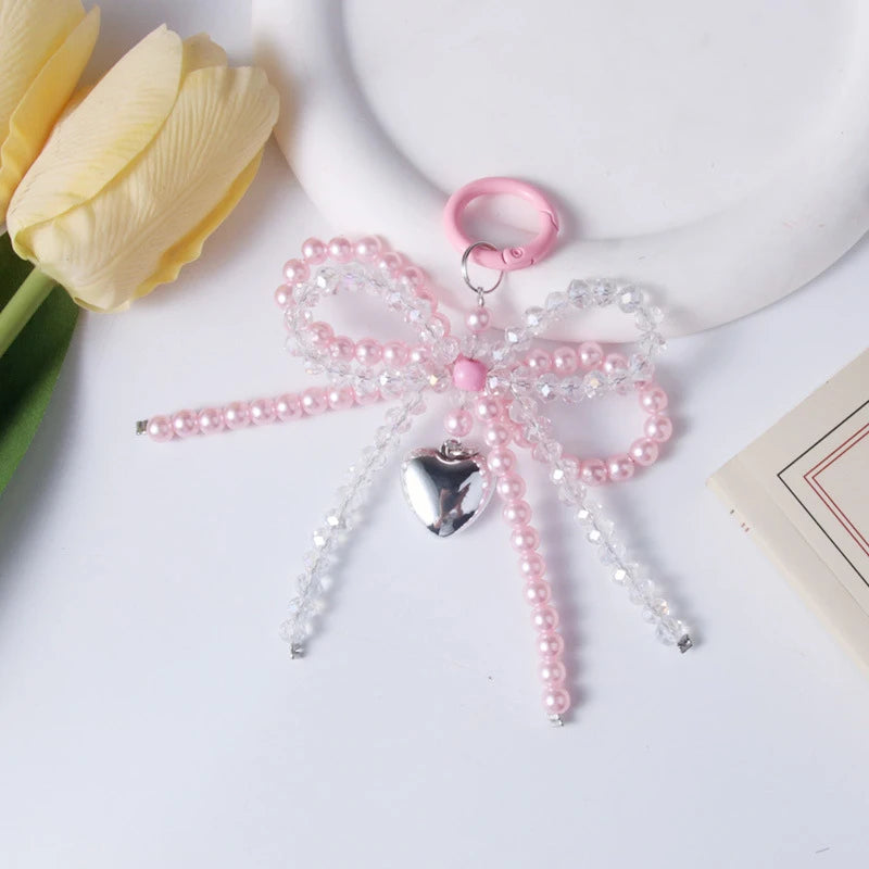 A pink pearl bowknot keychain featuring clear and pink beads and a silver heart charm, placed next to a yellow tulip.