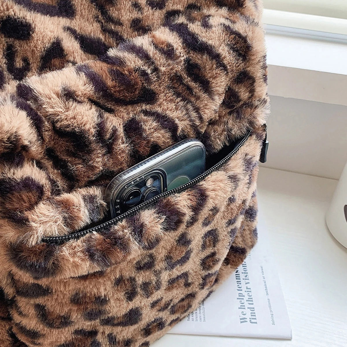 A close-up of a leopard print plush backpack with a zippered pocket showing a smartphone.