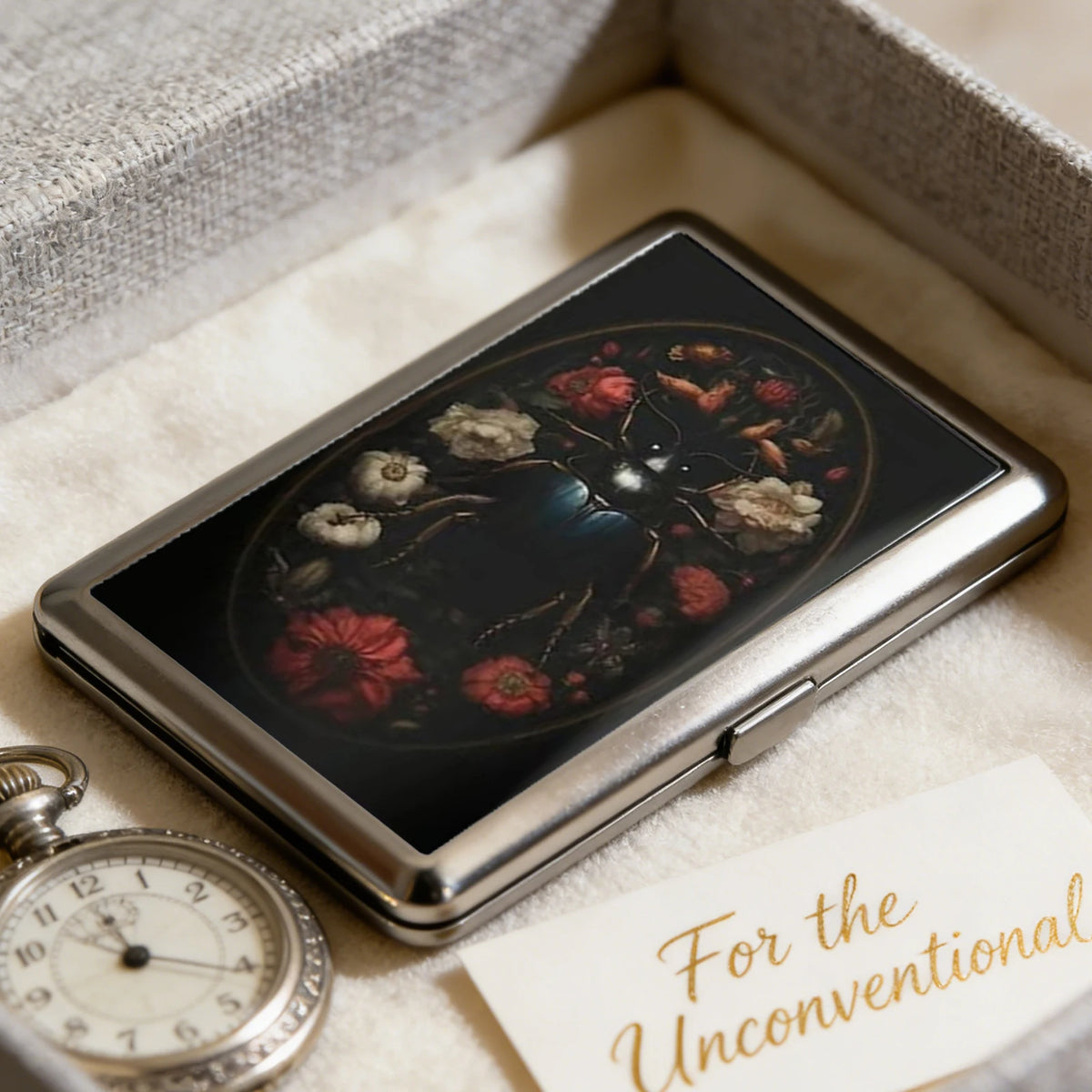 An open metal card case displaying a dark floral design on the lid, resting on a light fabric with a note.