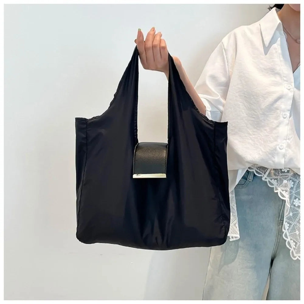 A person holds a black foldable mini keychain tote bag with a silver rectangular charm against a white background.