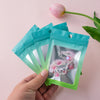 A hand holds a clear plastic bag filled with pink and white pearl jewelry inside a teal resealable storage bag.