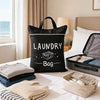 A black laundry bag with a handle sits on a white bed, surrounded by folded gray and blue towels.