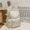 Mochila blanca con estampado floral y múltiples bolsillos, con un charm de oso blanco esponjoso sobre una superficie blanca.