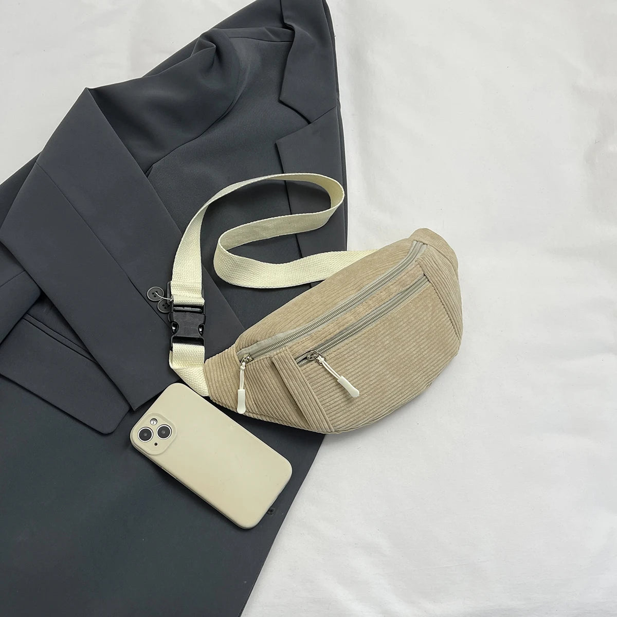 A beige corduroy crossbody waist bag with a light strap and a smartphone resting on a dark gray blazer.