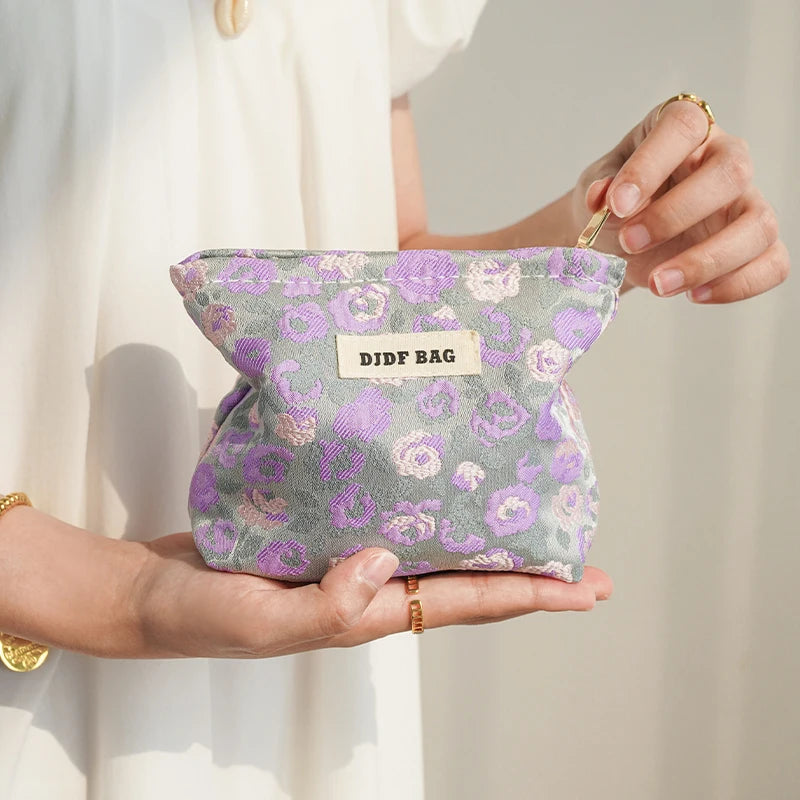 A purple floral canvas makeup bag with a gold charm is held in a person's hand against a light background.