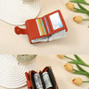 A brown leather compact wallet with a card slot and a zipper is shown next to a stack of cash and some dried flowers.