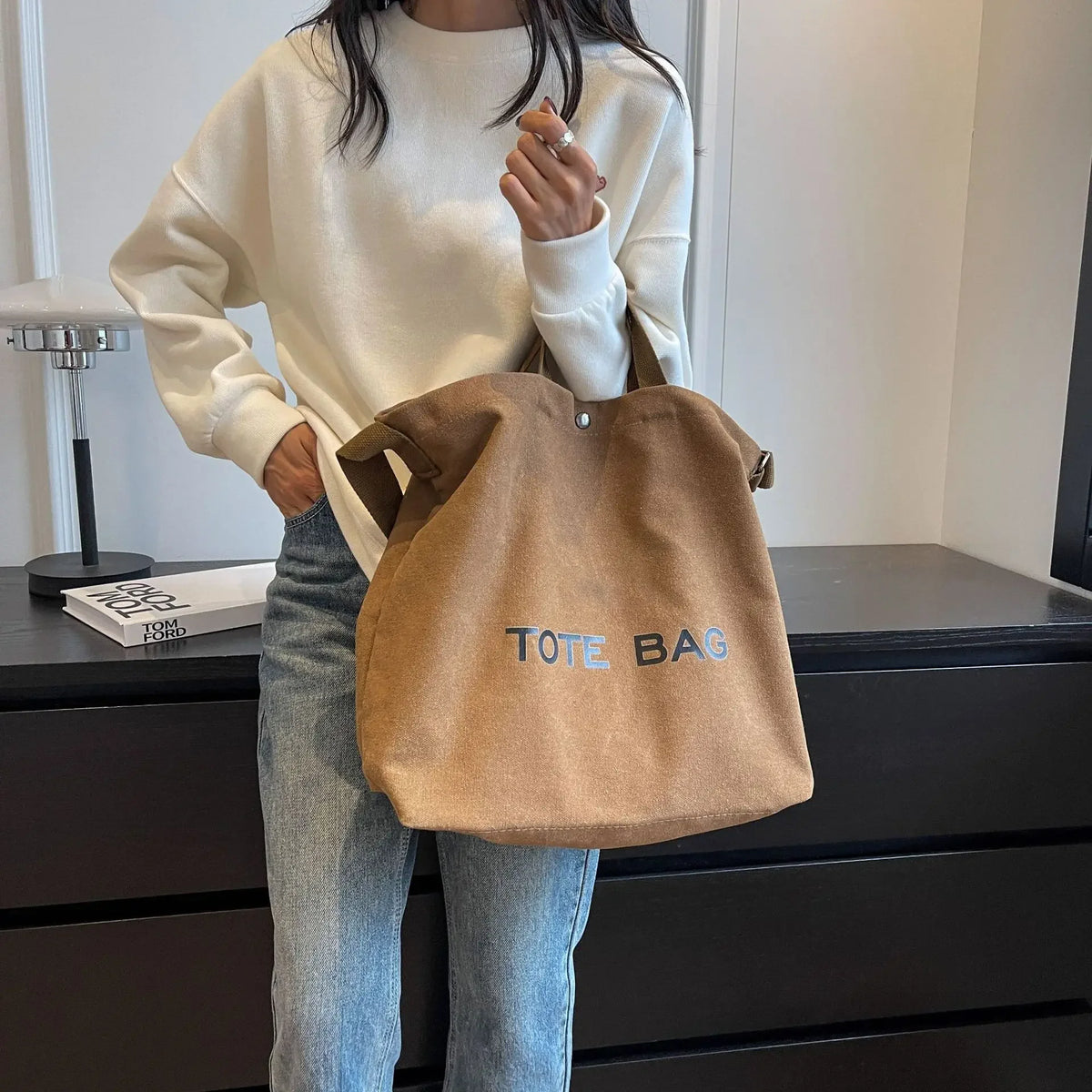 A woman wearing a cream sweater holds a brown canvas tote bag with blue lettering.
