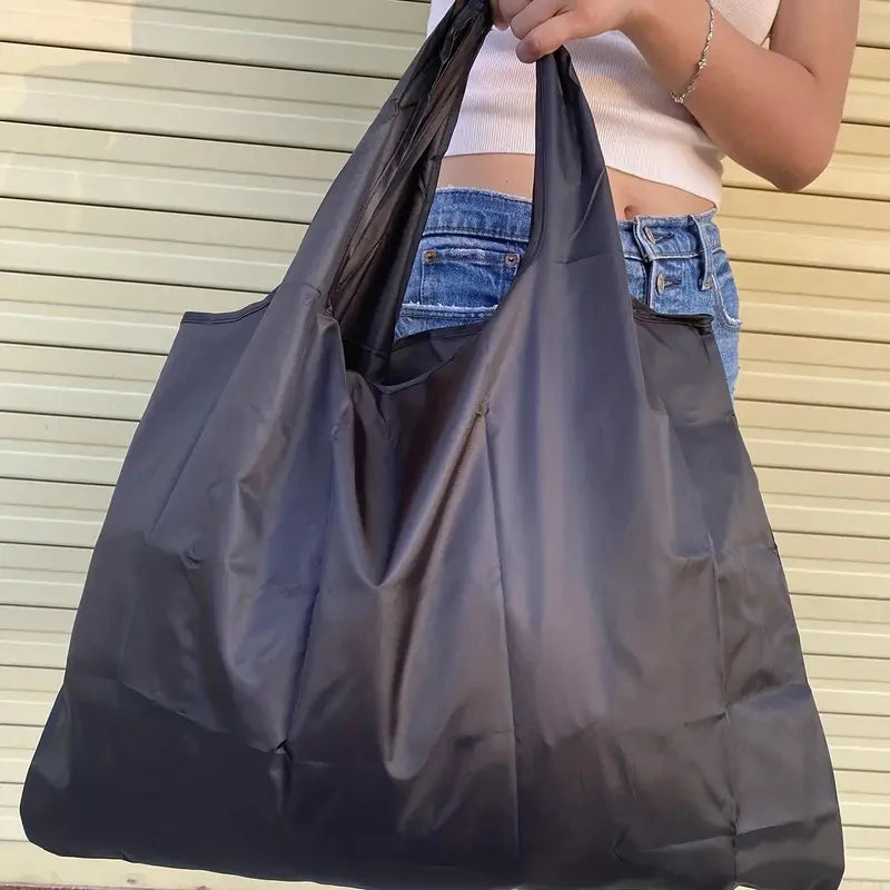 A person is holding a black foldable water-resistant shopping tote bag with a shoulder strap.