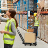 A worker in a safety vest is moving a cardboard box on a hand truck in a warehouse setting.