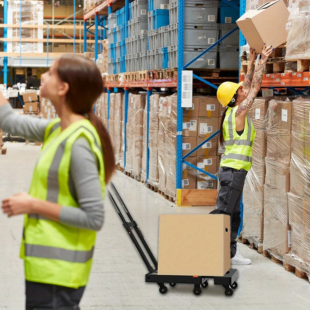 A worker in a safety vest is moving a cardboard box on a hand truck in a warehouse setting.