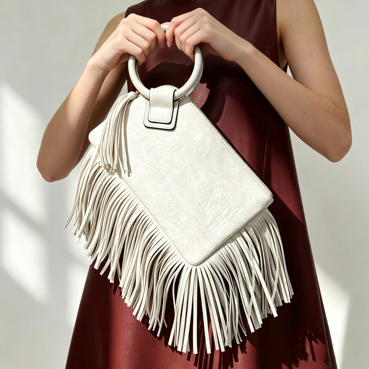 A woman holds a light beige fringed clutch bag with tassels against a dark red dress.