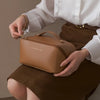 A woman holds a tan leather cosmetic storage bag against a brown skirt, showcasing the product.