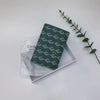 A dark green geometric print passport wallet resting on a white surface next to a leafy green branch.