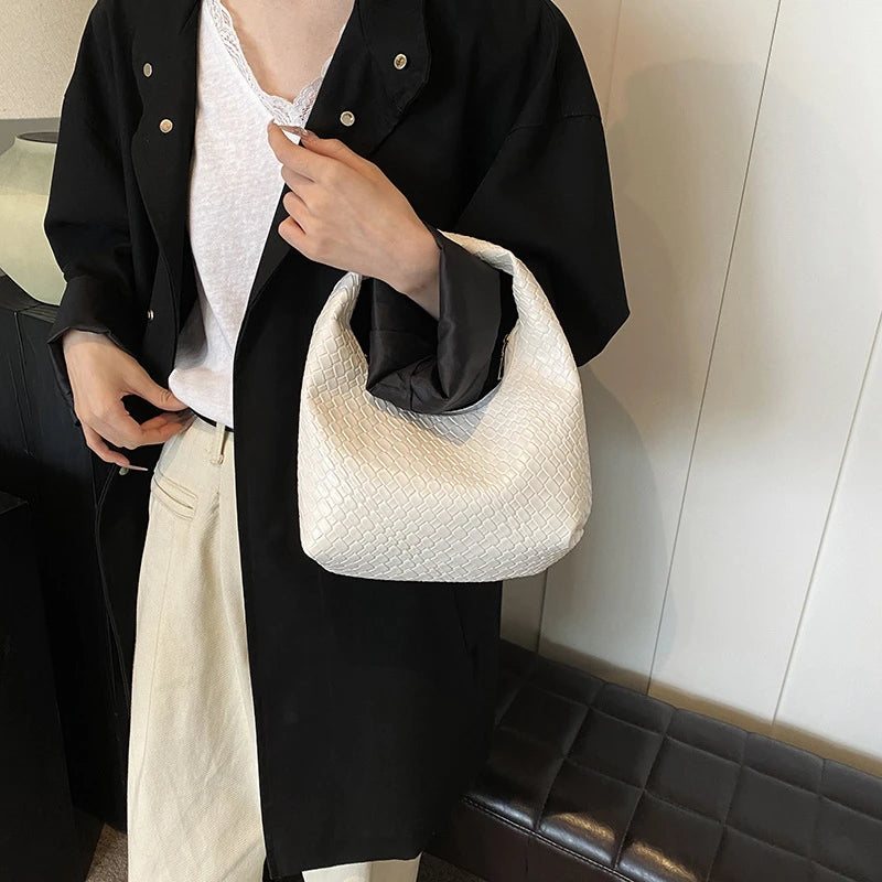 A person wearing a black shirt and light pants holds a cream woven embossed hobo shoulder bag with a top handle.