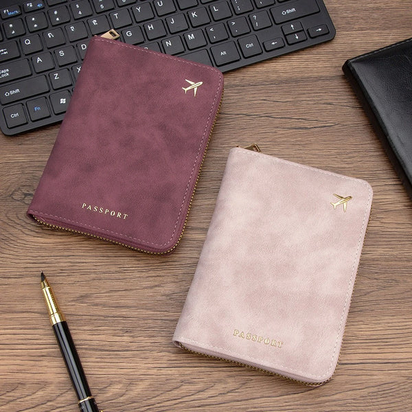 A light pink passport wallet with gold accents rests on a wooden desk next to a black laptop and a pen.