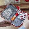 A hand opens a red compact survival and first aid kit with internal compartments and medical supplies.
