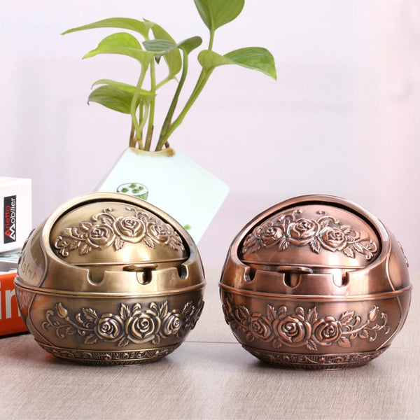A vintage metal ashtray with a rose embossing and windproof lid sitting on a wooden surface next to a potted plant.