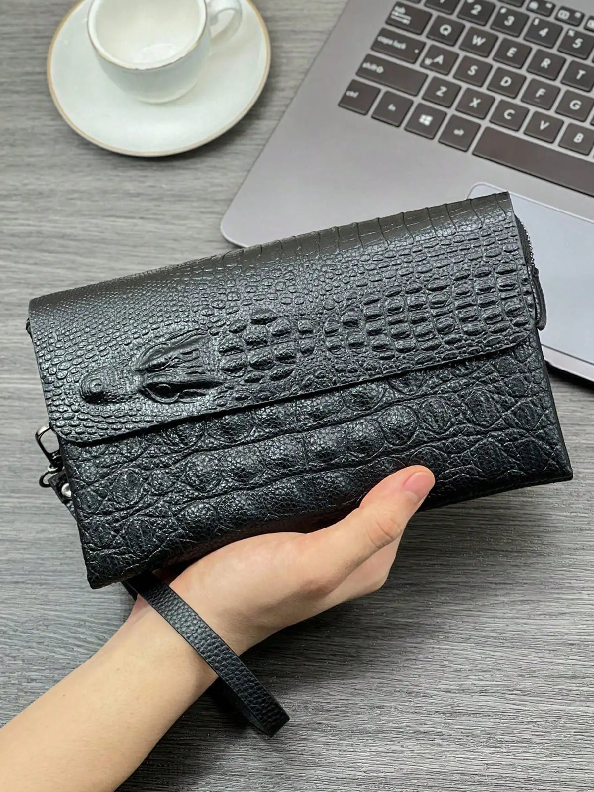 A hand holds a black crocodile pattern clutch bag next to a laptop and a cup on a gray surface.