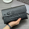 A hand holds a black crocodile pattern clutch bag next to a laptop and a cup on a gray surface.