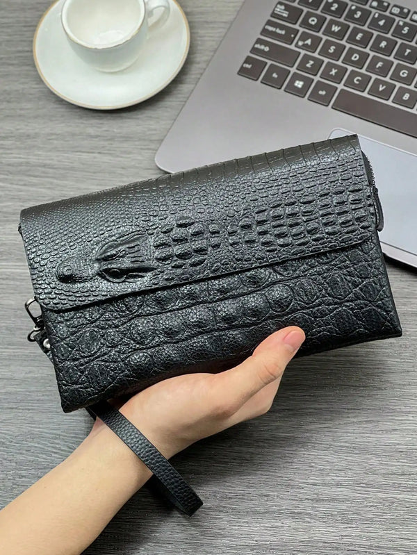 Eine Hand hält eine schwarze Krokodilmuster-Clutch neben einen Laptop und eine Tasse auf einer grauen Oberfläche.
