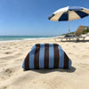 A navy blue and brown striped knit makeup bag resting on light sand near the ocean with a beach umbrella in the background.