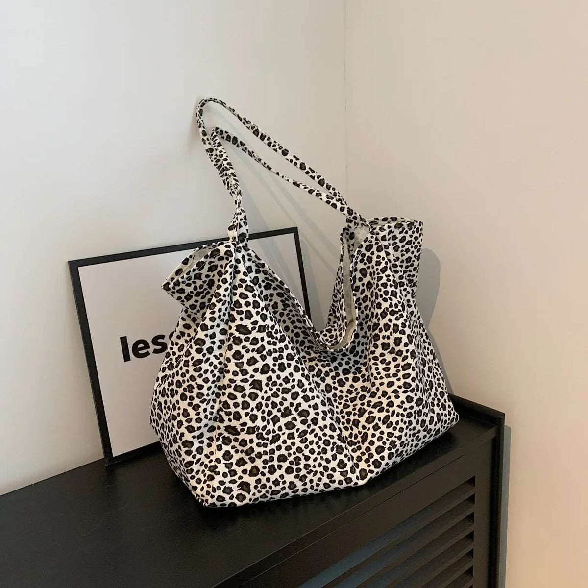 A leopard print canvas crossbody tote bag displayed on a black shelf with a framed sign.