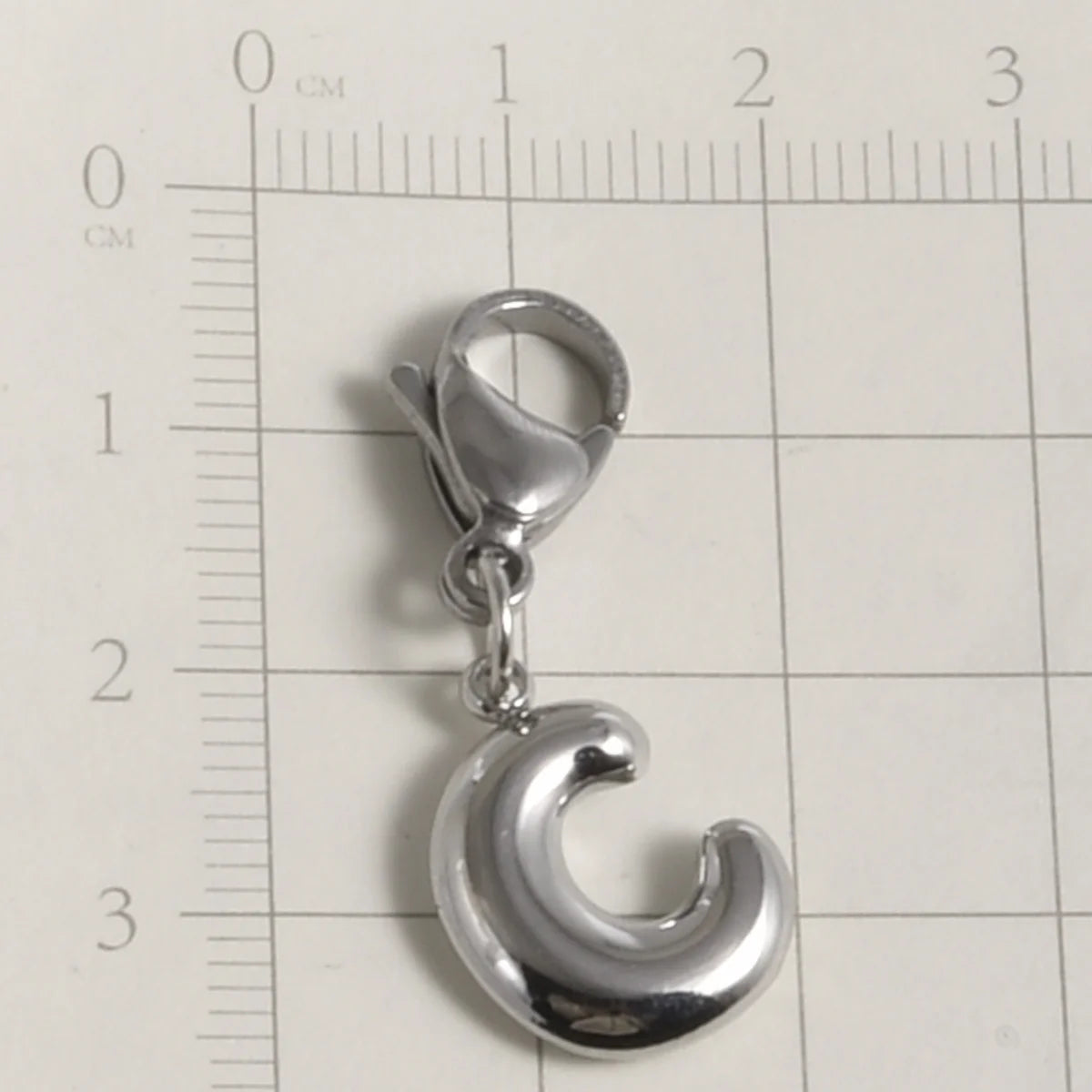 Silver stainless steel alphabet letter charm with a lobster clasp placed on a ruler.