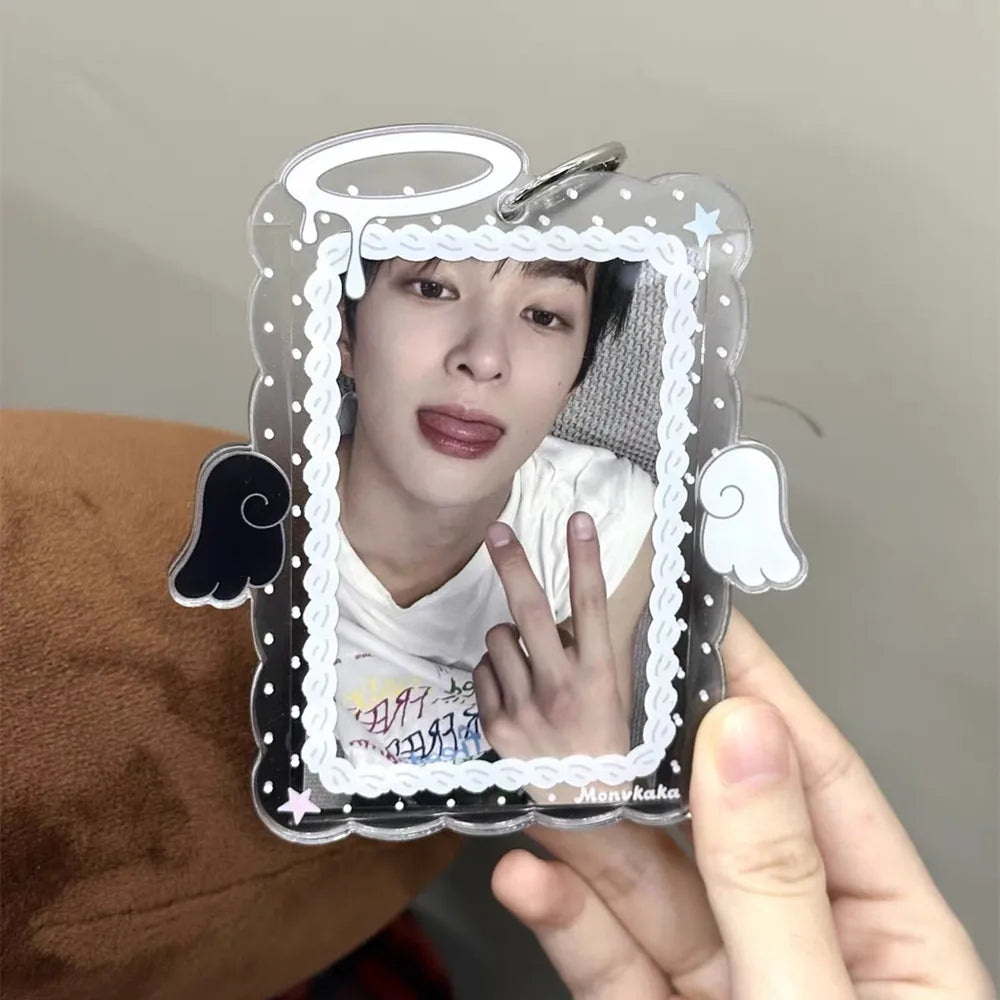 A hand holds a clear acrylic photocard holder with a cartoon angel wing design, featuring a person's face inside.