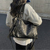 A woman is carrying a black leather tote bag with silver rivets and studs on the front, worn over a light gray sweatshirt.