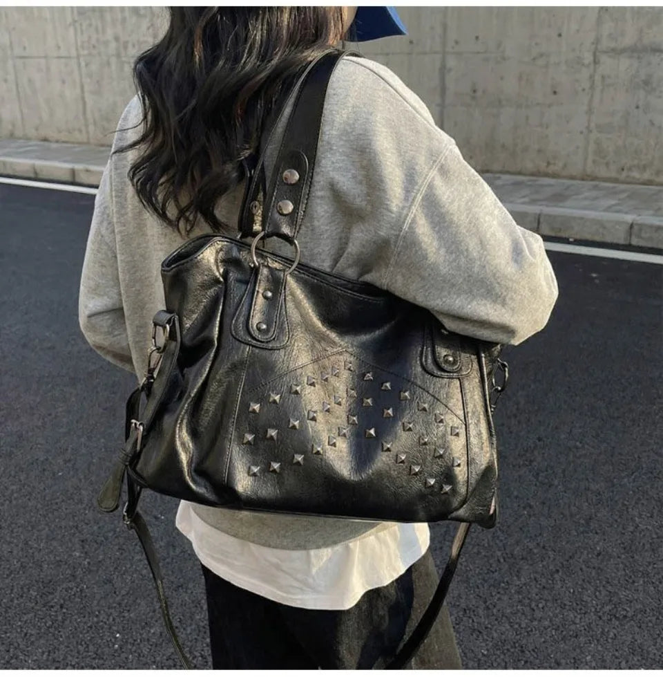 A woman is carrying a black leather tote bag with silver rivets and studs on the front, worn over a light gray sweatshirt.