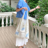 A woman wearing a blue top and white skirt holds a cream-colored knitted tote bag with blue embroidery on a porch.