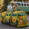 A quilted travel duffle bag decorated with colorful floral and nature patterns, resting on asphalt near a vintage van.