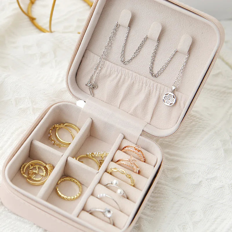 A light pink compact leather jewelry travel box with velvet lining displaying silver and gold rings and necklaces.
