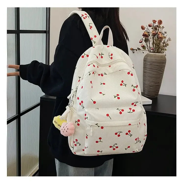 A white corduroy fashion backpack with a red cherry pattern and pink accents is shown against a dark background.
