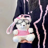 A pink and white plush bear keychain wearing a camera and glasses is held against a dark patterned fabric.