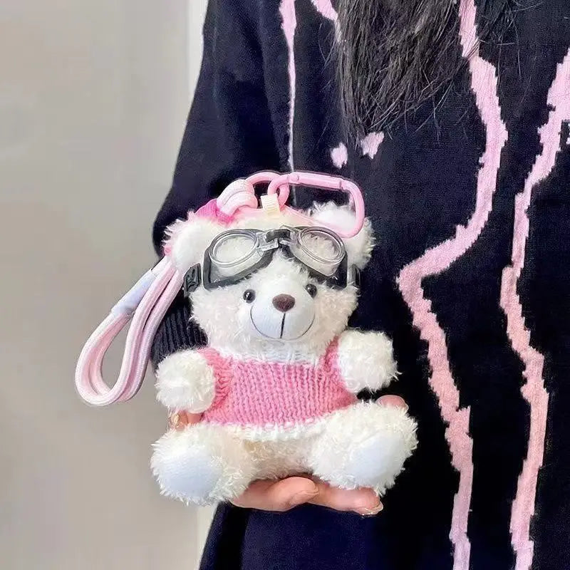 A pink and white plush bear keychain wearing a camera and glasses is held against a dark patterned fabric.