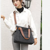 A woman wearing a white shirt and gray pants holds a black canvas tote bag with brown straps against a light wall.
