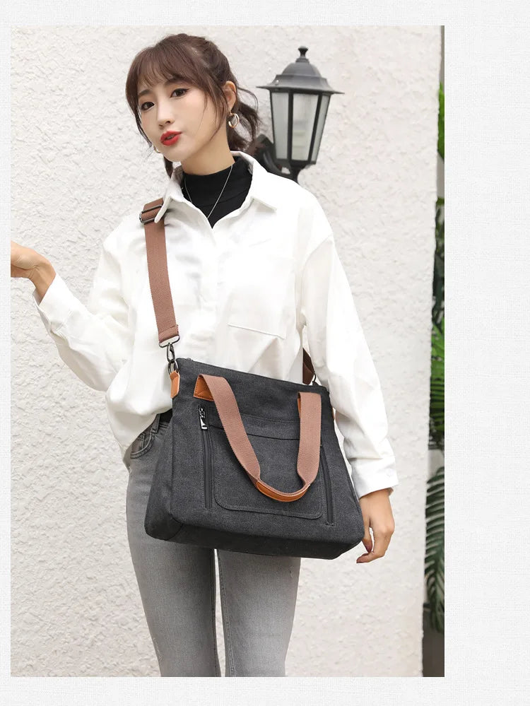 A woman wearing a white shirt and gray pants holds a black canvas tote bag with brown straps against a light wall.