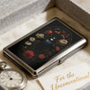 A silver metal card holder with a dark background featuring an intricate floral design resting in a light-colored box.