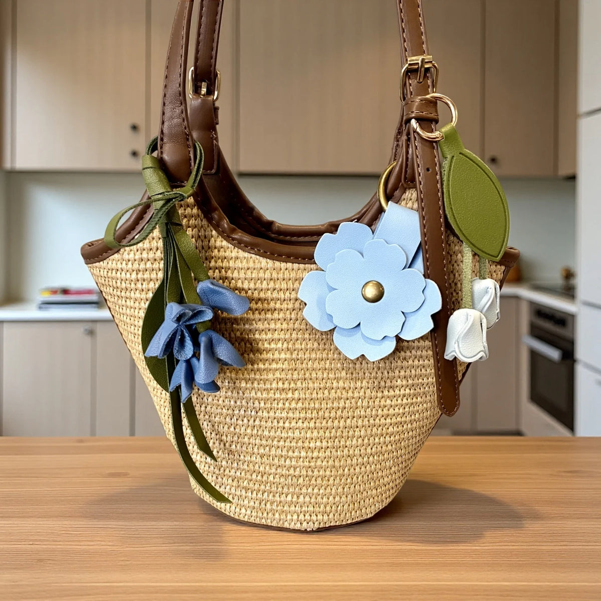 A tan woven leather shoulder bag with brown straps and blue floral charms resting on a wooden table.